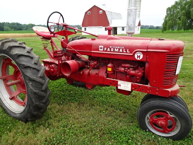 Röd veterantraktor av märket Farmall på en gräsmatta framför en lada.